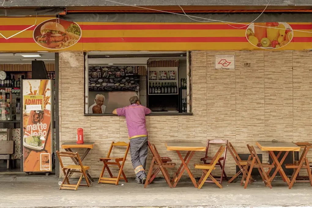 SEO Local para restaurantes. Uma pessoa vestindo uma camisa rosa está parada em frente a uma lanchonete de rua com mesas e cadeiras de madeira.