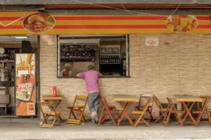 SEO Local para restaurantes. Uma pessoa vestindo uma camisa rosa está parada em frente a uma lanchonete de rua com mesas e cadeiras de madeira.